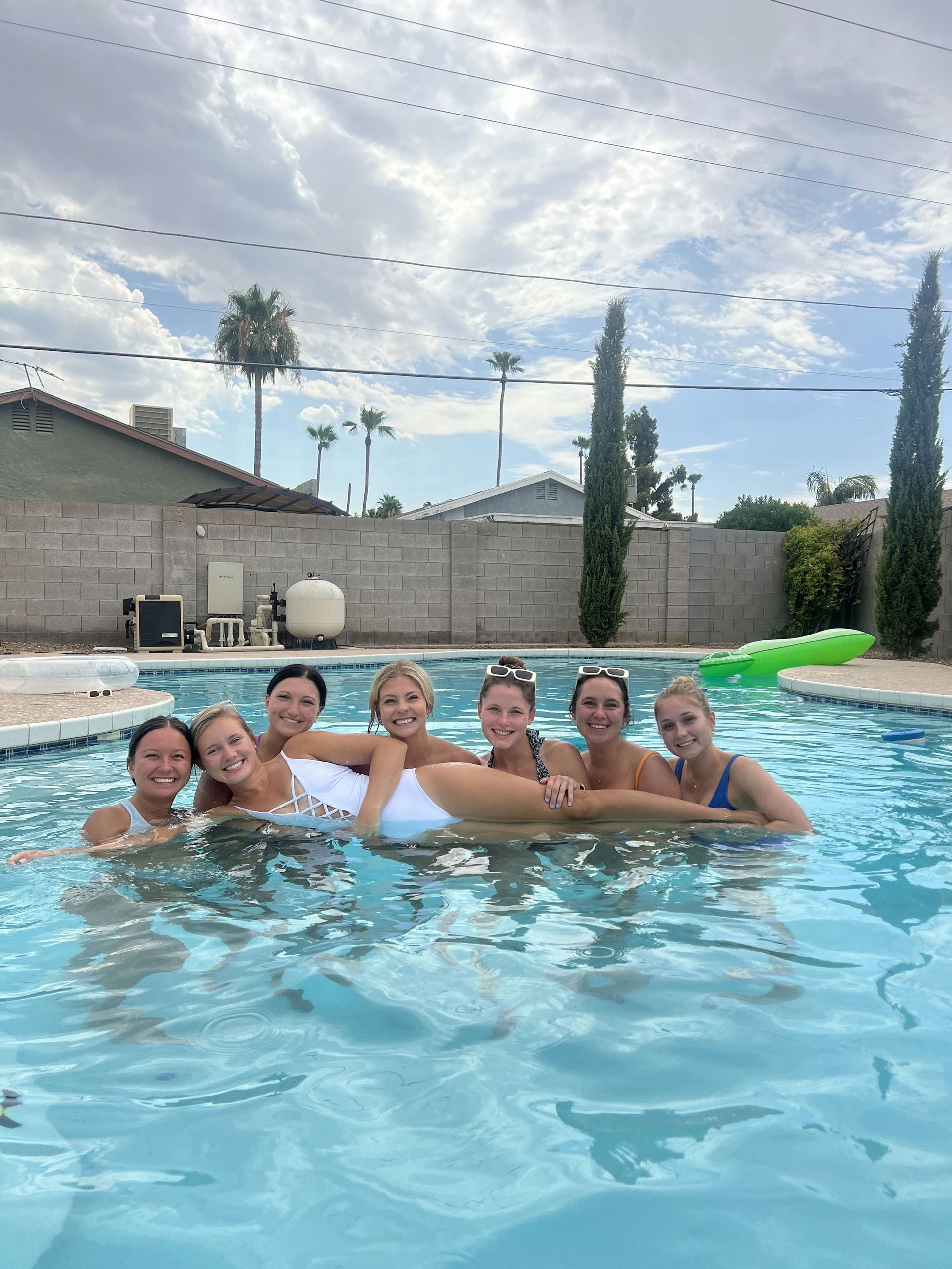 Seven friends in swimsuits smiling in a backyard pool while holding one friend horizontally above the water, with palm trees, a cinderblock wall and a green inflatable raft under a partly cloudy sky.