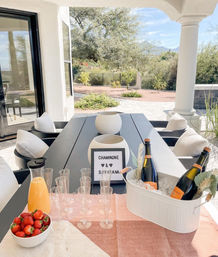 Covered outdoor patio dining table staged for a champagne brunch—ice bucket with champagne bottles, plastic flutes, orange juice carafe and bowl of strawberries, overlooking a desert backyard with cacti and distant mountains.