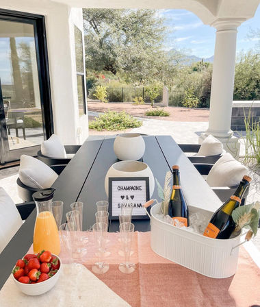 Covered outdoor patio dining table staged for a champagne brunch—ice bucket with champagne bottles, plastic flutes, orange juice carafe and bowl of strawberries, overlooking a desert backyard with cacti and distant mountains.