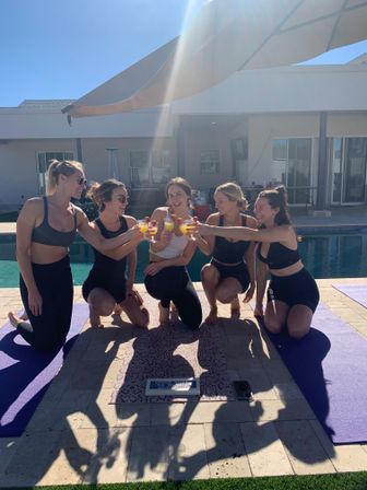 Five women in workout clothes kneeling on yoga mats by a sunny backyard pool, smiling and toasting mimosas under a patio umbrella — poolside yoga brunch vibe.