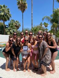 Twelve women in swimsuits smiling and posing by a sunny backyard pool framed by tall palm trees and a clear blue sky, one holding a small letterboard—festive bachelorette pool party vibe.