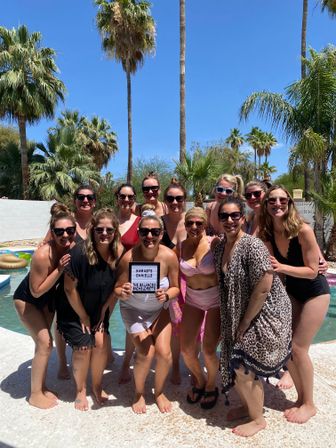 Twelve women in swimsuits smiling and posing by a sunny backyard pool framed by tall palm trees and a clear blue sky, one holding a small letterboard—festive bachelorette pool party vibe.