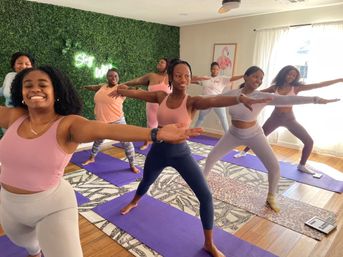 Group of women in a bright home yoga studio practicing Warrior II on purple mats, smiling in front of a faux green plant wall with neon sign and flamingo wall art.