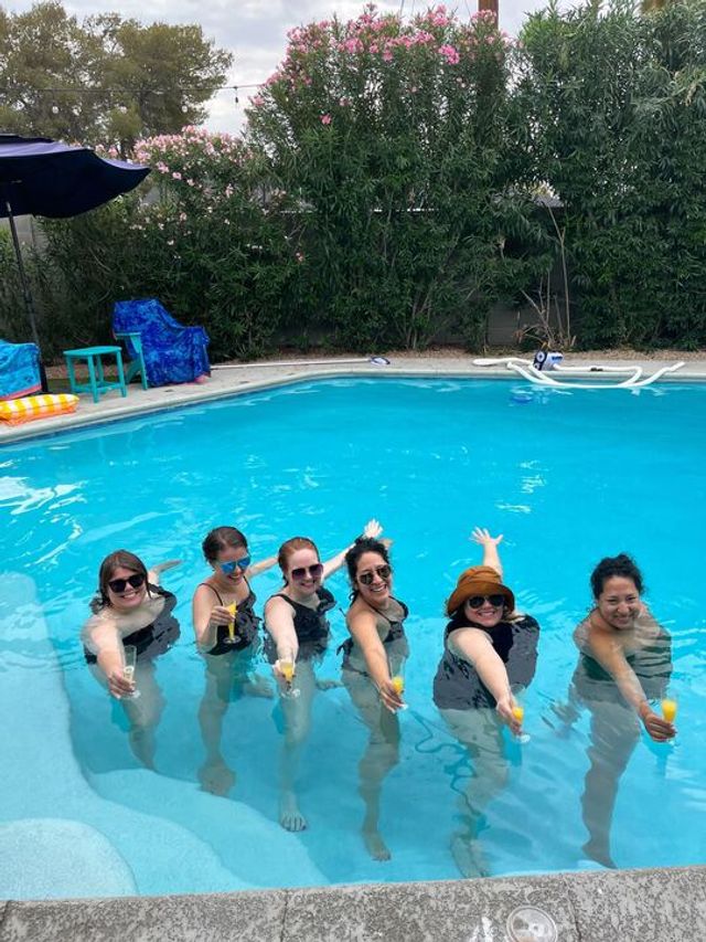 Backyard pool party with six women standing in bright blue water, each holding a mimosa, pool umbrella and colorful chairs on the deck, pink-flowering hedge backdrop