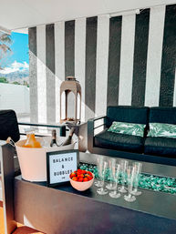 Sunlit modern outdoor patio with black-and-white vertical striped wall, dark cushioned sofa with tropical leaf pillows, ice bucket of champagne and juice, bowl of strawberries, six champagne flutes and turquoise glass fire pit, with mountains visible beyond a white courtyard wall.