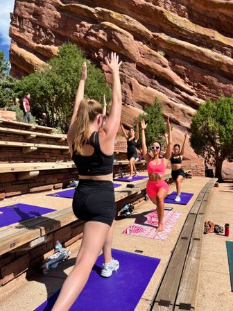 Sunlit outdoor yoga class on red-rock amphitheater steps — women on colorful mats in lunges with arms raised against towering red sandstone cliffs and juniper trees.