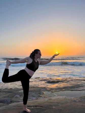 Person doing dancer yoga pose barefoot on a rocky beach at sunset, silhouetted against a warm orange sky and ocean waves, playfully appearing to hold the sun.
