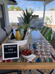 Sunny backyard patio table setup under a pergola with an ice bucket holding champagne and juice, bowl of strawberries, rose-gold champagne flutes, potted agave centerpiece and a letter board reading 'CHAMPAGNE AND SAVASANA' on a colorful woven runner.