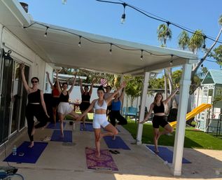 Group of women practicing tree pose on yoga mats during an outdoor class under a covered patio in a sunny backyard with palm trees, string lights and a children's playset.