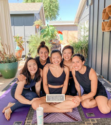 Five friends in black activewear smiling on purple yoga mats in a sunny backyard patio, posing for a bachelorette-themed outdoor yoga photo surrounded by potted plants.