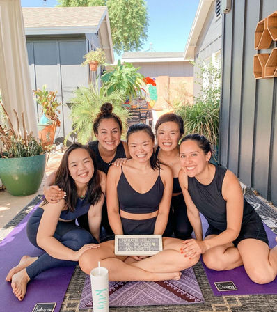 Five friends in black activewear smiling on purple yoga mats in a sunny backyard patio, posing for a bachelorette-themed outdoor yoga photo surrounded by potted plants.