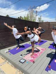 Outdoor yoga class on a sunny wooden deck — women in playful balancing poses on purple and patterned mats, smiling and reaching forward with water bottles and sunglasses nearby