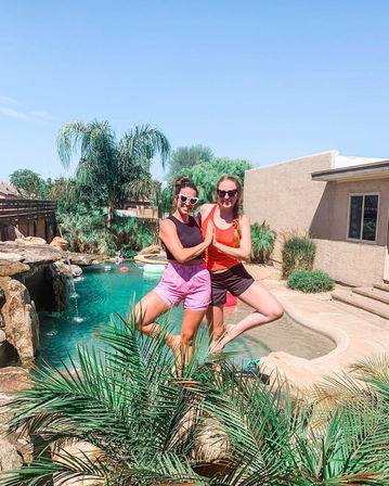Two friends in heart-shaped sunglasses doing tree-pose by a turquoise backyard pool with palm trees, rock waterfall and stucco house on a sunny summer day.