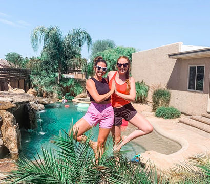 Two friends in heart-shaped sunglasses doing tree-pose by a turquoise backyard pool with palm trees, rock waterfall and stucco house on a sunny summer day.