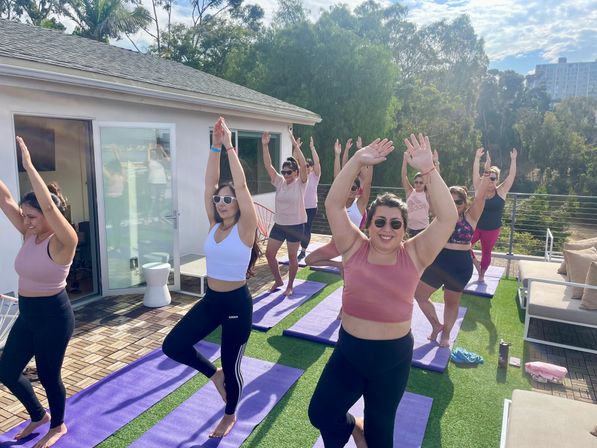 Outdoor rooftop yoga class of women doing tree pose on purple mats on a sunny terrace with trees and distant buildings.
