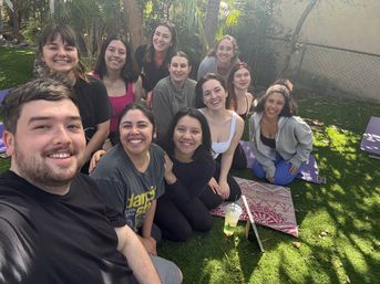 Smiling group selfie of a dozen adults on yoga mats in a sunny backyard — outdoor yoga/community wellness session on a green lawn with trees.
