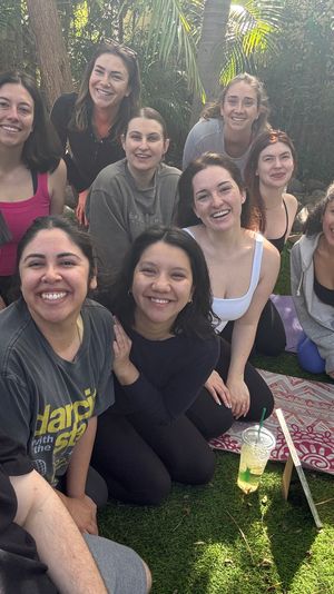 Smiling group selfie of a dozen adults on yoga mats in a sunny backyard — outdoor yoga/community wellness session on a green lawn with trees.