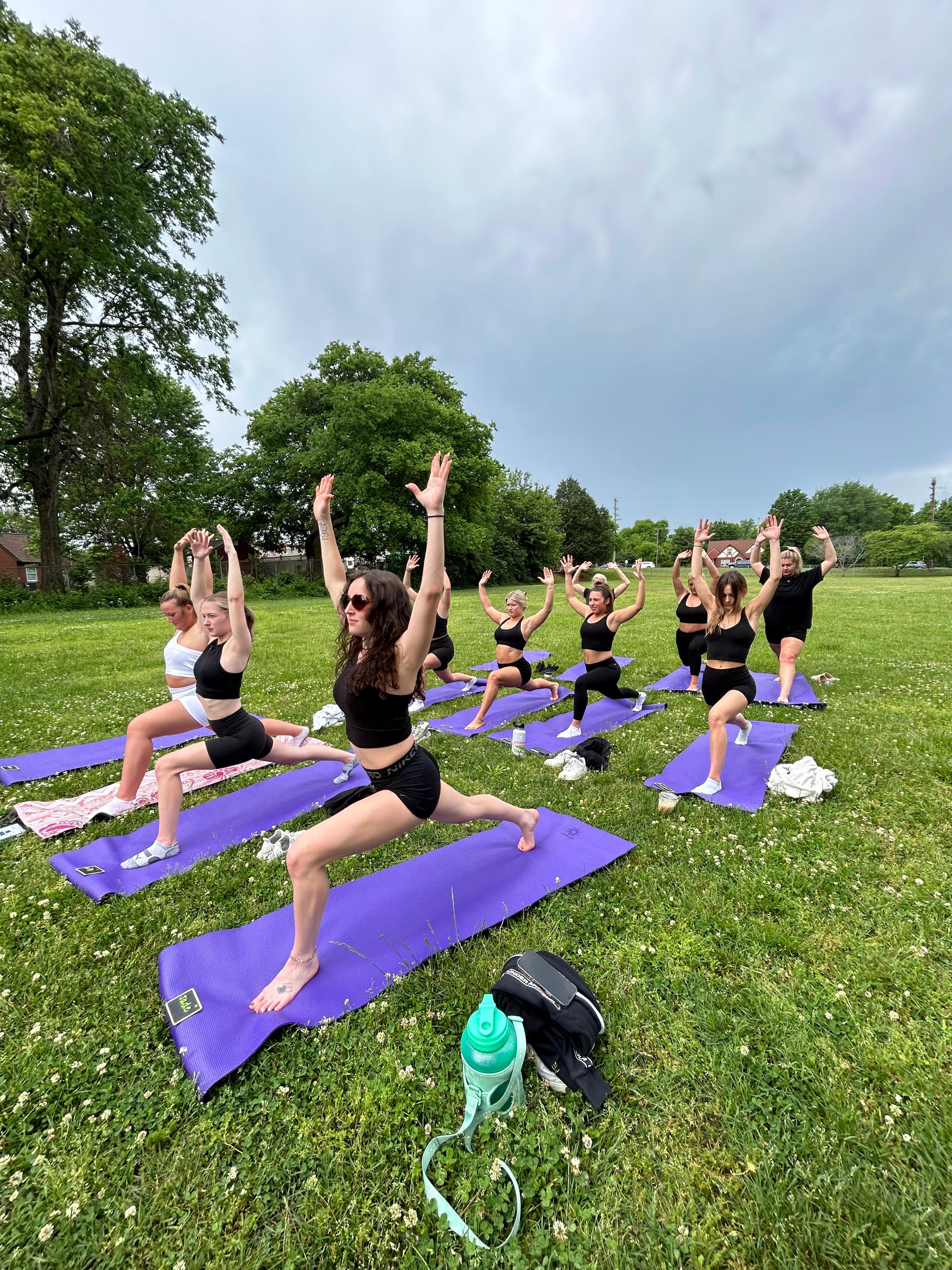 Outdoor group yoga class in a green park with women on purple mats doing high lunge/warrior poses under a cloudy sky