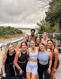 Cheerful group of nine women in activewear posing on a lakeside deck with trees, shoreline and an overcast sky — scenic outdoor fitness meetup photo.