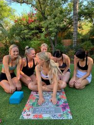 Six women in activewear laughing during a bridal outdoor yoga session on grass with a colorful yoga mat and block, Scottsdale, AZ.
