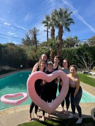Group of women in athletic wear posing by a backyard pool under palm trees, holding a large pink heart-shaped inflatable float on a sunny blue-sky day