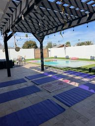 Sunlit backyard pool patio with black wooden pergola and string lights, purple yoga mats laid on tiled floor beside a pool with inflatable rings and palm trees in the background