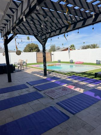 Sunlit backyard pool patio with black wooden pergola and string lights, purple yoga mats laid on tiled floor beside a pool with inflatable rings and palm trees in the background
