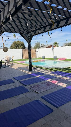 Sunlit backyard pool patio with black wooden pergola and string lights, purple yoga mats laid on tiled floor beside a pool with inflatable rings and palm trees in the background