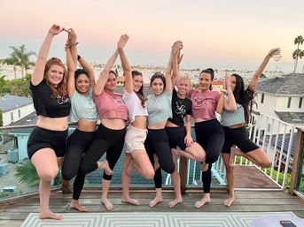 Eight friends in crop tops and leggings striking tree-pose on a waterfront balcony at sunset with a marina and palm trees in the background, one holding a wine glass — playful group celebration.