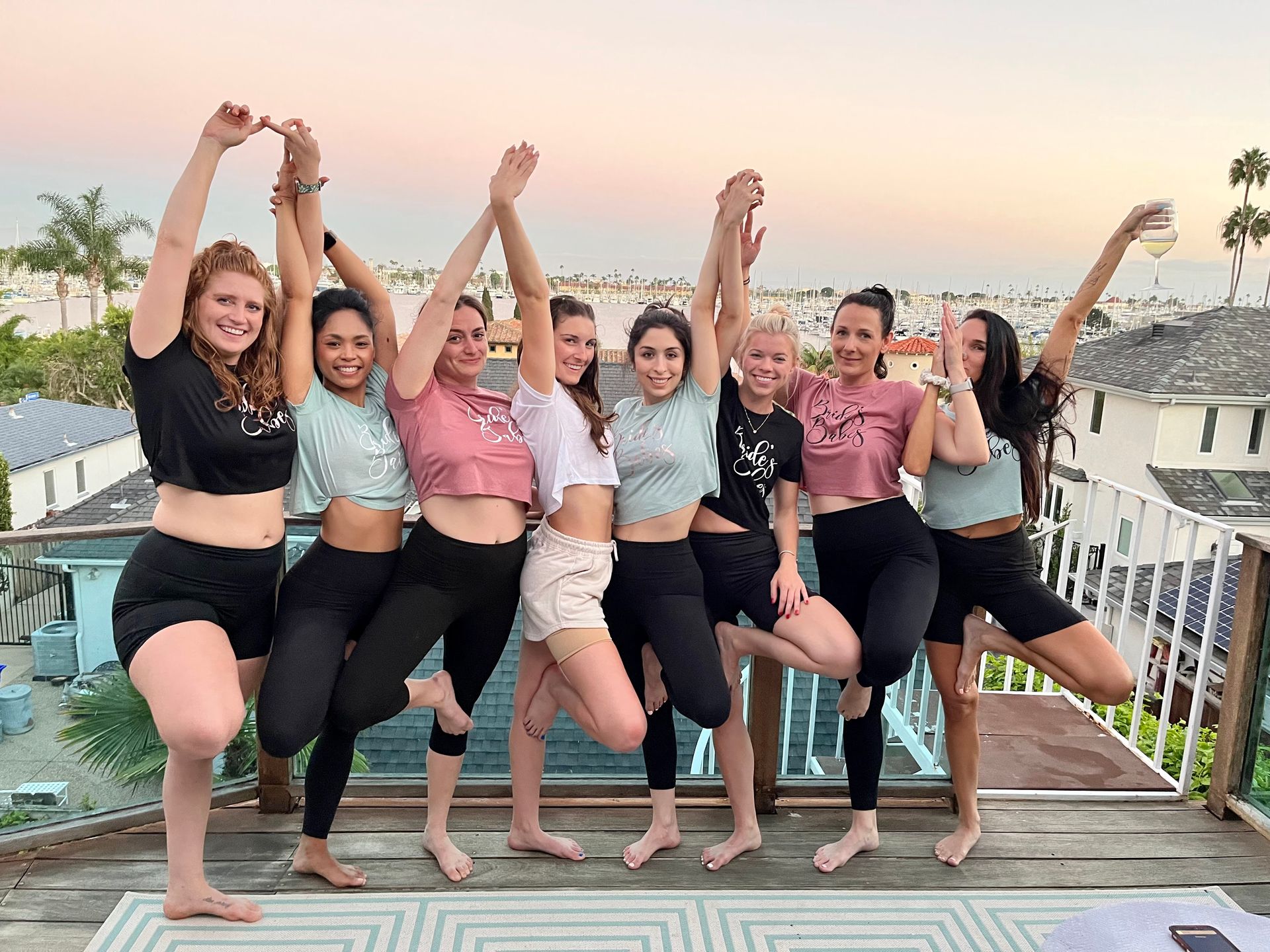 Eight friends in crop tops and leggings striking tree-pose on a waterfront balcony at sunset with a marina and palm trees in the background, one holding a wine glass — playful group celebration.