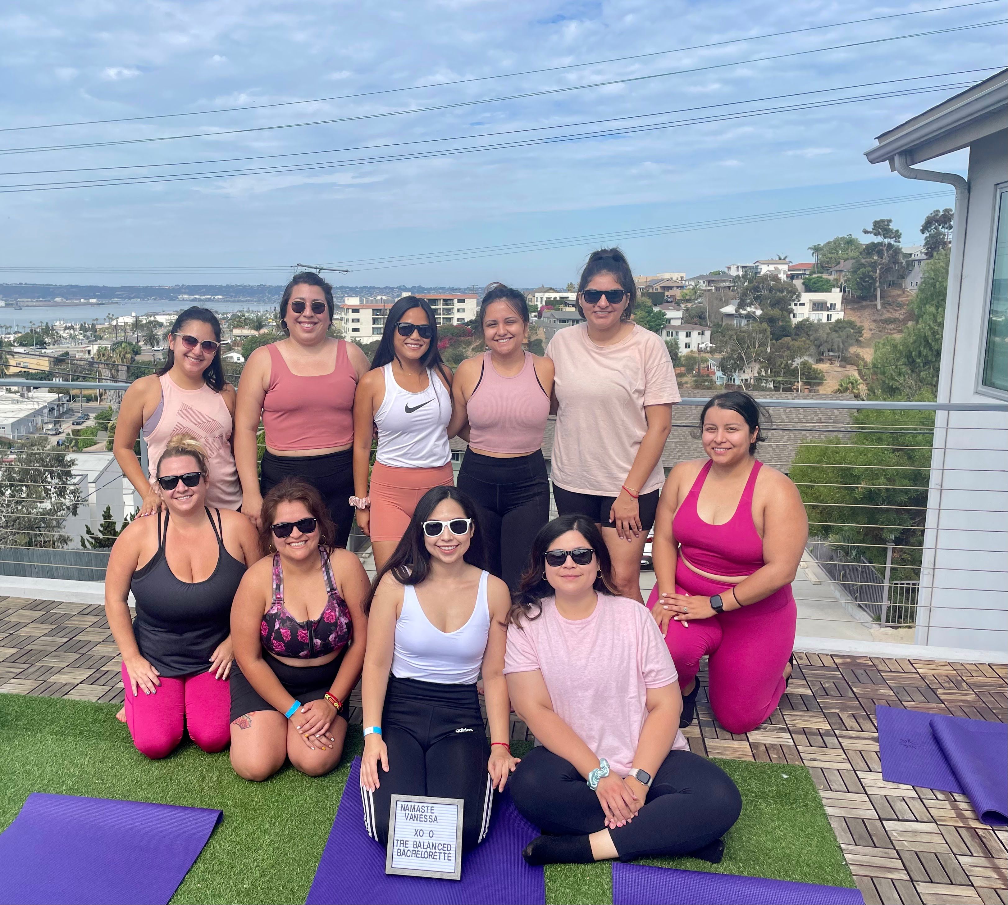 Rooftop yoga group of ten women in colorful activewear smiling on purple mats with a coastal bay and hillside homes in the background