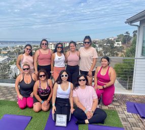 Rooftop yoga group of ten women in colorful activewear smiling on purple mats with a coastal bay and hillside homes in the background