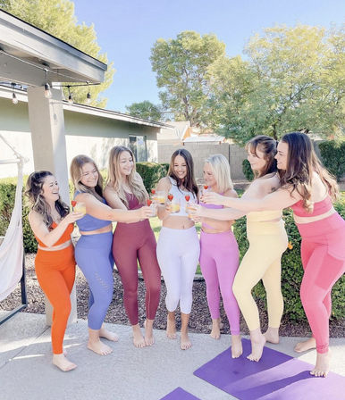 Seven friends in colorful workout leggings and crop tops toast with strawberry-garnished orange drinks in a sunny backyard, barefoot on yoga mats.