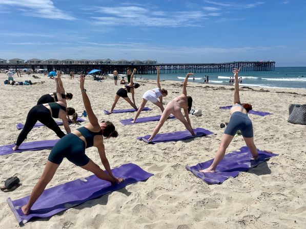 Group beach yoga class on purple mats doing triangle stretches on a sunny sandy shoreline with ocean waves and a wooden pier under a blue sky