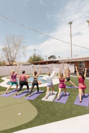 Group yoga class with seven people on purple mats in a sunny suburban backyard beside a kidney-shaped pool, performing high lunge poses on artificial turf under a blue sky with tall palm trees.