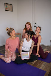 Four smiling women in casual activewear sitting on purple yoga mats in a bright indoor yoga studio, laughing together and holding a small bachelorette sign — playful group yoga session.
