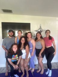 Seven friends in activewear posing on purple yoga mats in a bright living room with silver star balloons and a wall-mounted TV, smiling after a home yoga session.