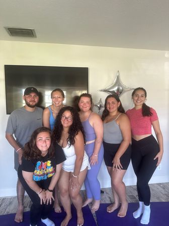 Seven friends in activewear posing on purple yoga mats in a bright living room with silver star balloons and a wall-mounted TV, smiling after a home yoga session.