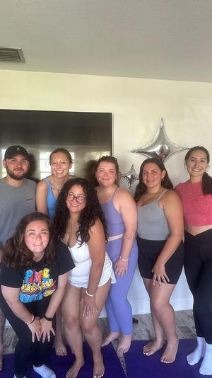 Seven friends in activewear posing on purple yoga mats in a bright living room with silver star balloons and a wall-mounted TV, smiling after a home yoga session.