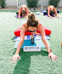 Sunny outdoor yoga class on green turf — woman in red leggings in extended child's pose reaching forward while sipping from a canned drink on her mat, with two other students stretching on mats behind her.