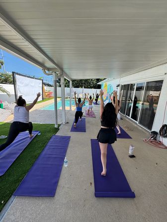 Outdoor poolside yoga class on a covered patio, students on purple mats doing lunge/warrior poses facing a residential swimming pool and colorful mural