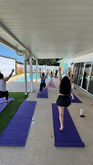 Outdoor poolside yoga class on a covered patio, students on purple mats doing lunge/warrior poses facing a residential swimming pool and colorful mural