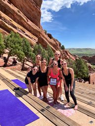 Six women in heart-shaped sunglasses pose together on wooden tiered seating at a red-rock outdoor amphitheater in Colorado, smiling on yoga mats during a sunny bachelorette yoga outing with canyon and blue sky in the background.