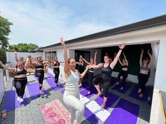 Outdoor rooftop yoga class on a sunlit terrace with an instructor and a group of women smiling while balancing in tree pose on purple mats