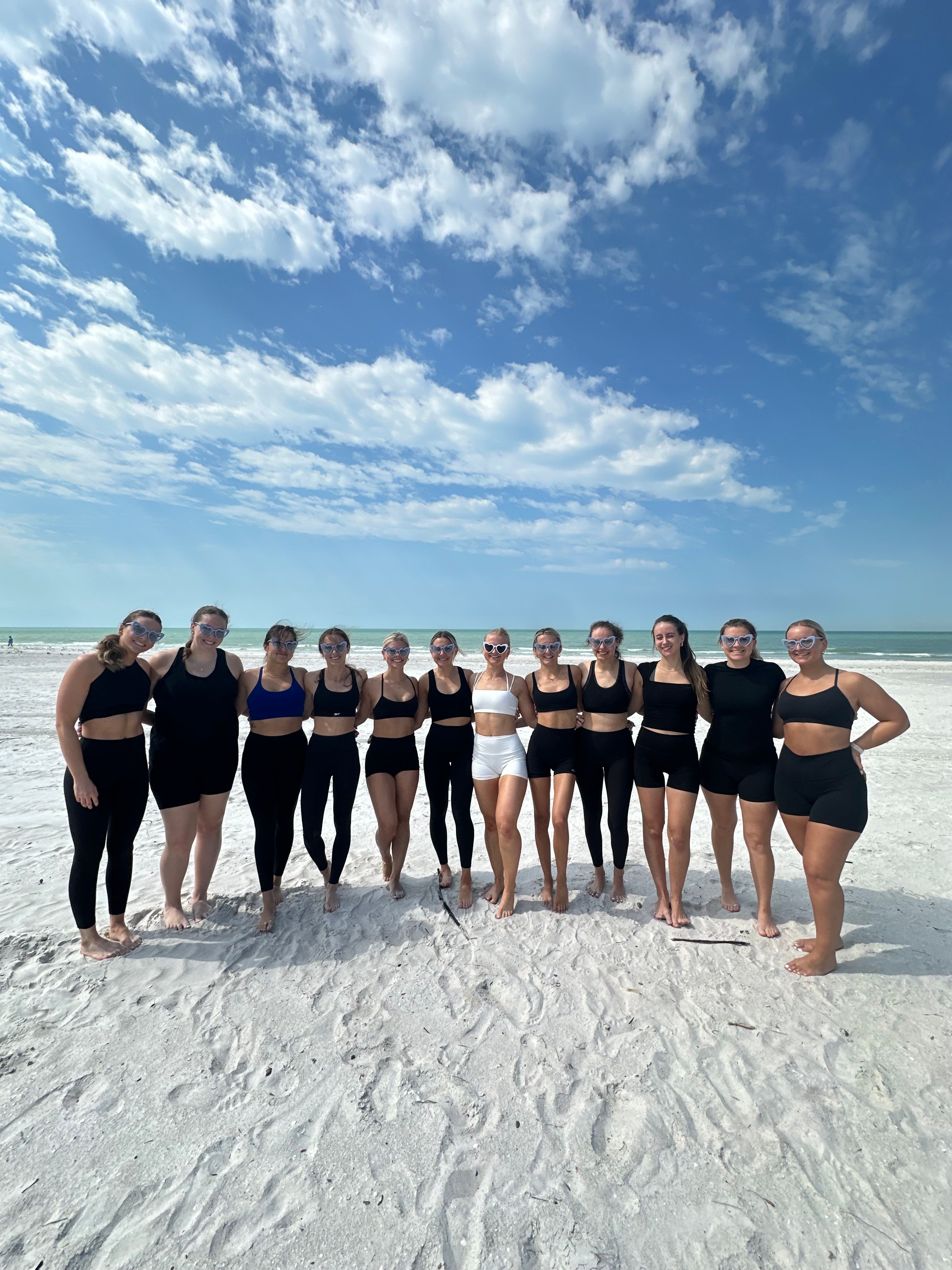 Group of 13 women in black and white athletic swimwear and sunglasses posing on a white-sand beach with turquoise ocean and a bright blue, cloud-dotted sky.