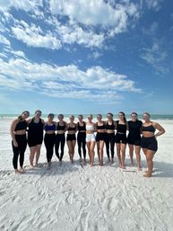 Group of 13 women in black and white athletic swimwear and sunglasses posing on a white-sand beach with turquoise ocean and a bright blue, cloud-dotted sky.