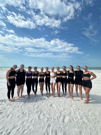 Group of 13 women in black and white athletic swimwear and sunglasses posing on a white-sand beach with turquoise ocean and a bright blue, cloud-dotted sky.