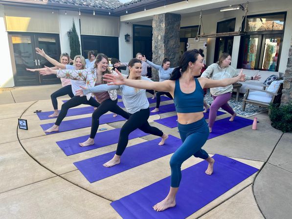 Women’s outdoor yoga class on a sunny backyard patio — group in Warrior II on purple mats, smiling and stretching during a casual wellness session.