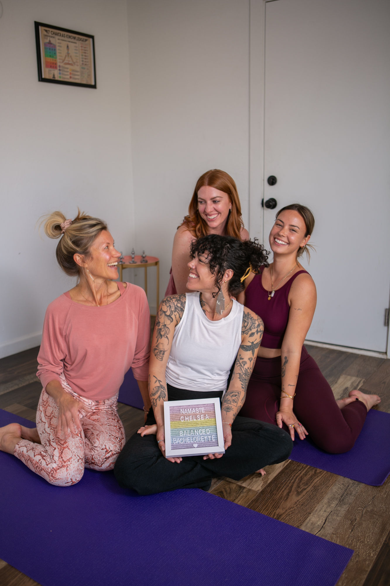 Four smiling women in casual activewear sitting on purple yoga mats in a bright indoor yoga studio, laughing together and holding a small bachelorette sign — playful group yoga session.