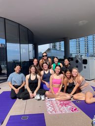 Group of women smiling on yoga mats during a sunny rooftop yoga session with glass high-rises and city skyline in the background.